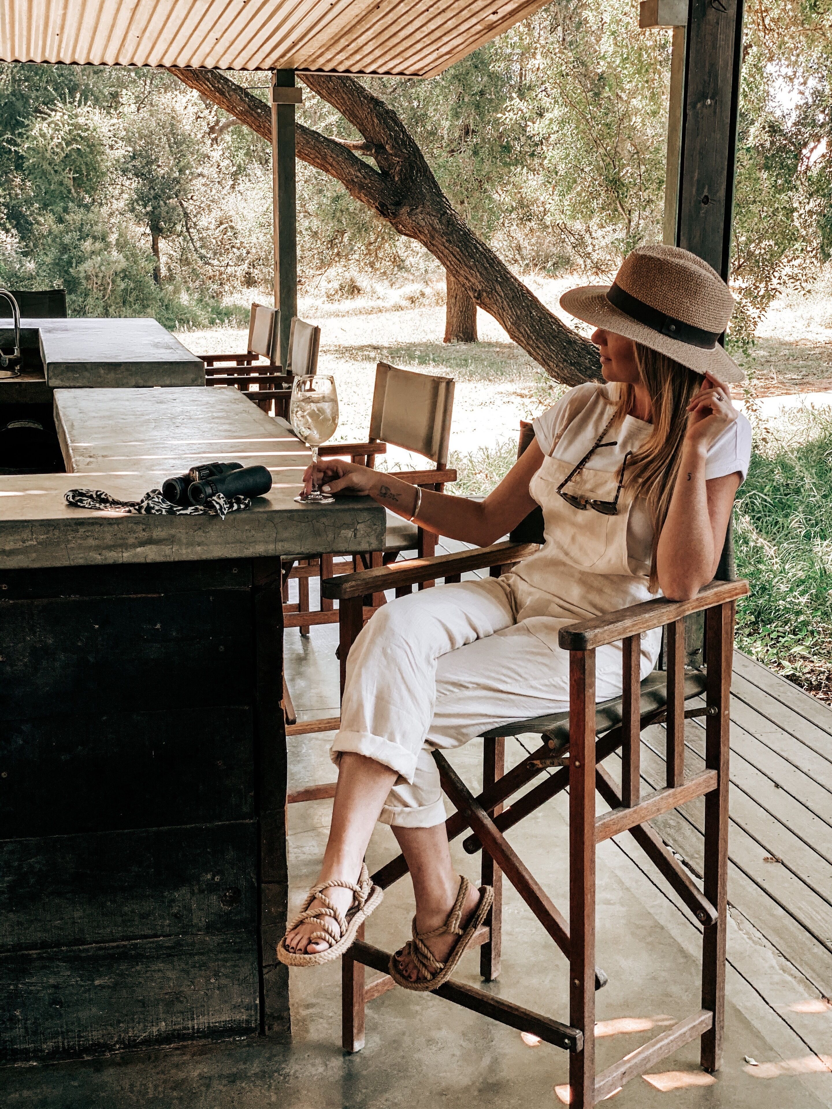 A person seated at a wooden table with a wine glass and binoculars, under a shaded area with a view of trees.