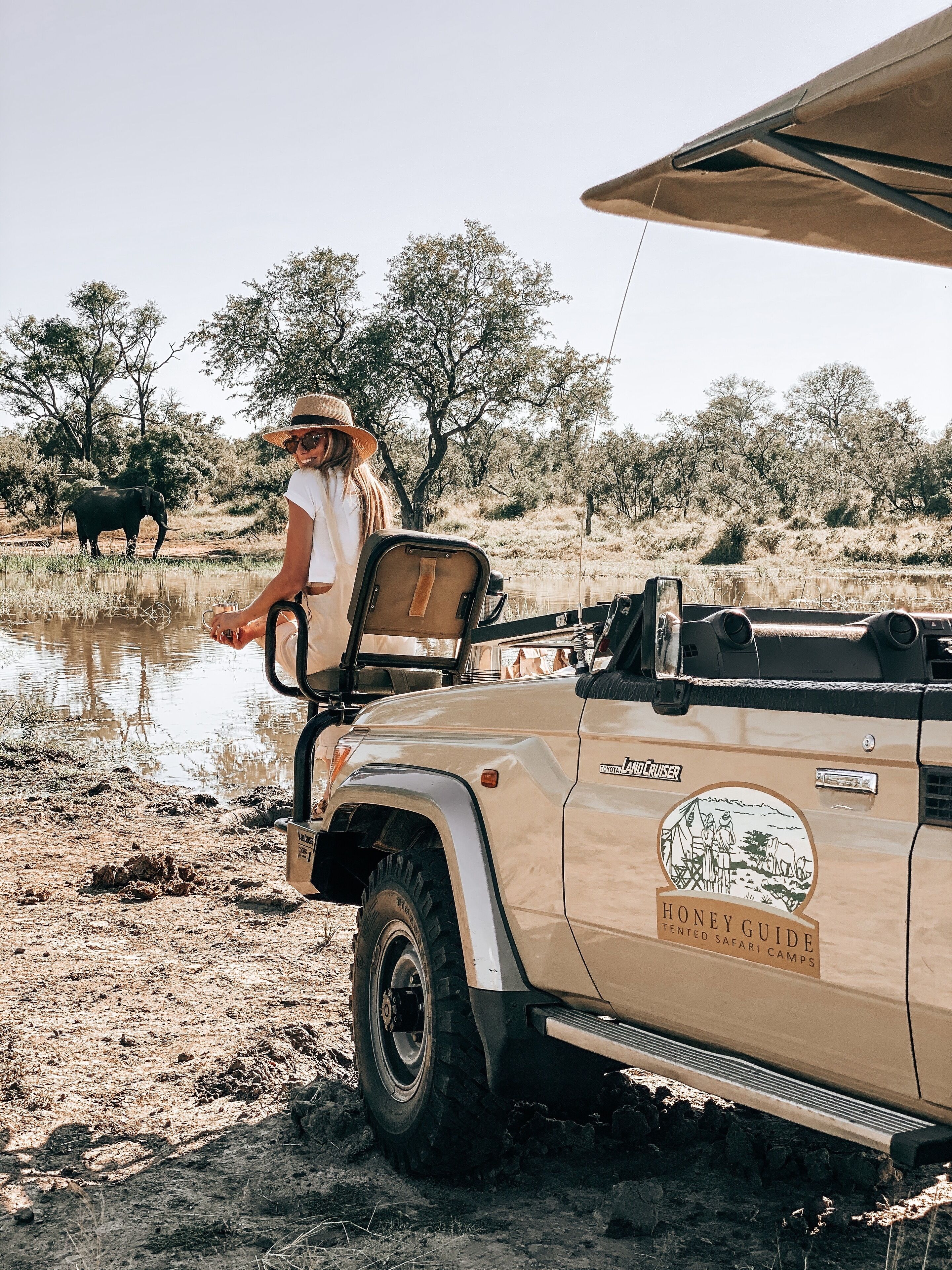 A safari vehicle with a "Toyota Land Cruiser" and "Honey Guide Tented Safari Camps" logo, parked near a water body with trees and an elephant in the background.