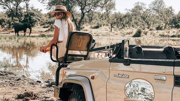 A safari vehicle with a "Toyota Land Cruiser" and "Honey Guide Tented Safari Camps" logo, parked near a water body with trees and an elephant in the background.
