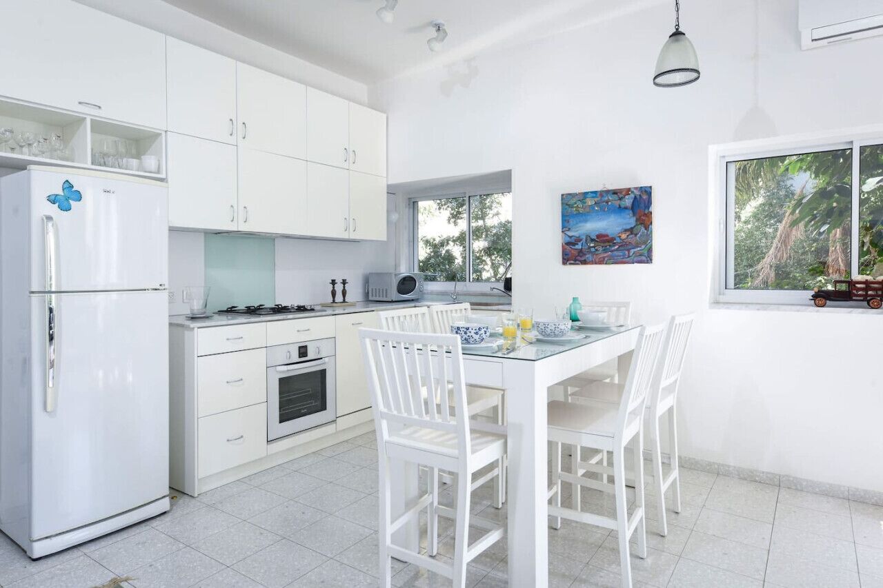 A modern kitchen with white cabinetry, a dining table with chairs, a refrigerator, and a window with a view of trees.