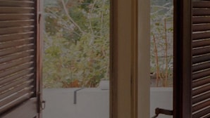 A view through a window with wooden shutters, revealing a garden with trees and a white wall. - Tranquil Garden Retreat by Sea N Rent (Tel Aviv)