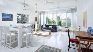 A spacious living area with a dining table, white chairs, a grey sofa, and a large window with curtains. - Tranquil Garden Retreat by Sea N Rent (Tel Aviv)