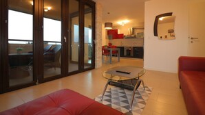 Ein modernes Wohnzimmer mit einer Glastür, die auf einen Balkon führt, einem roten Sofa, einem Glastisch und einer Küche im Hintergrund.