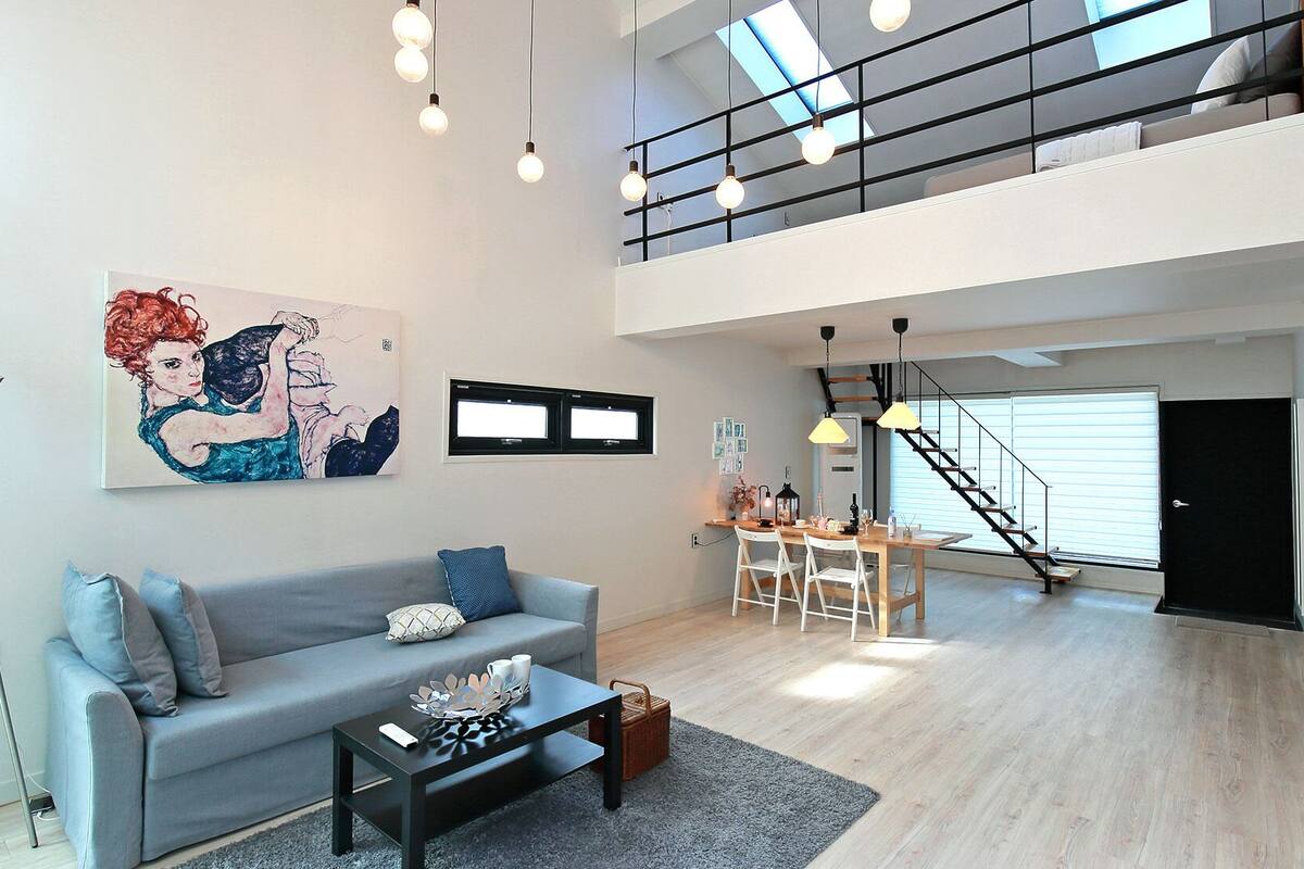 Een ruime woonkamer met een grijze bank, een eettafel en een trap die naar een hoger gelegen verdieping leidt.