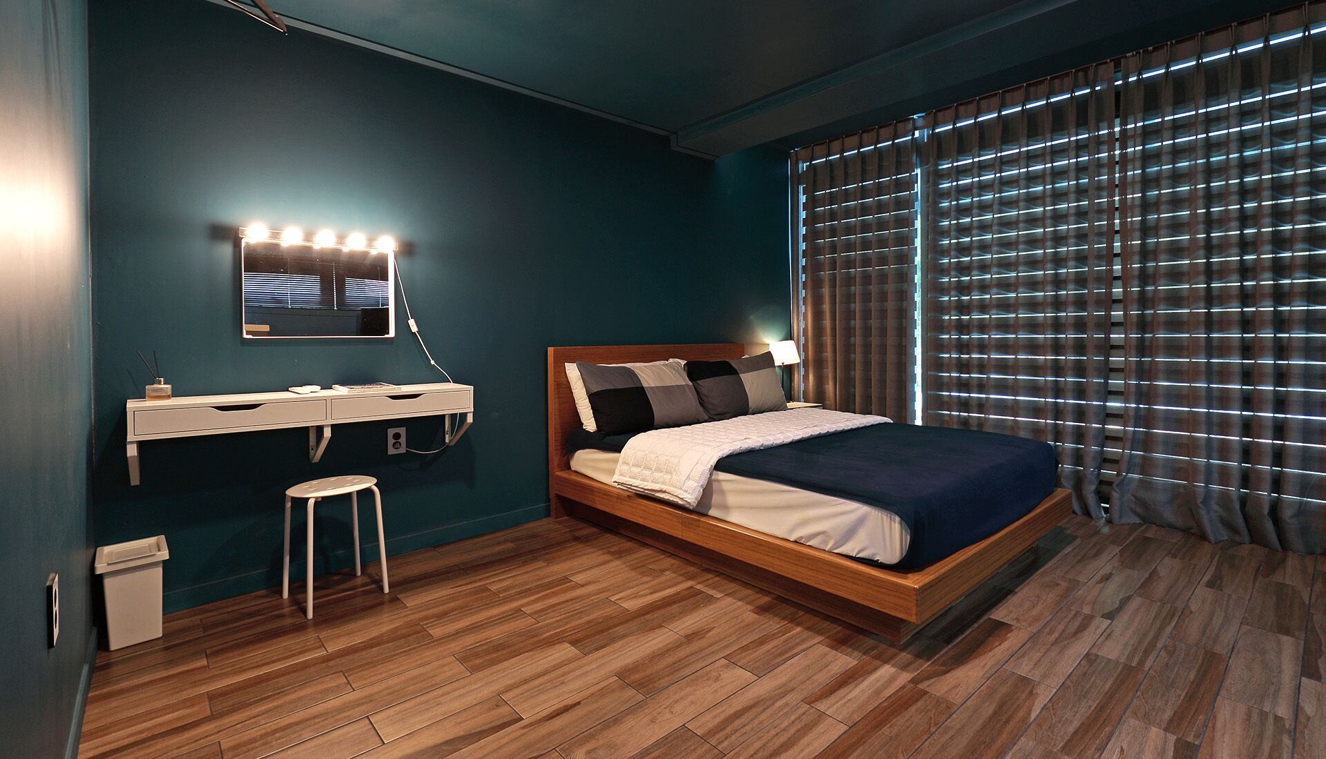 A bedroom with a wooden bed, a vanity with a mirror and lights, a small stool, and a window with blinds.
