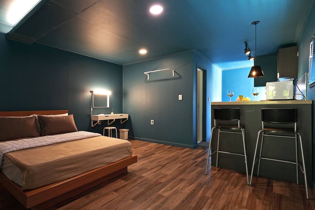 Een moderne slaapkamer met een groot bed, nachtkastjes, een bureau met stoel, en een kitchenette.
