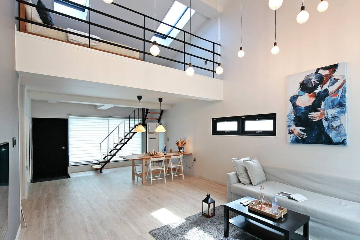 Een ruime woonkamer met een hoge plafond, een trap naar een hoger niveau, een eethoek en een groot abstract schilderij aan de muur.
