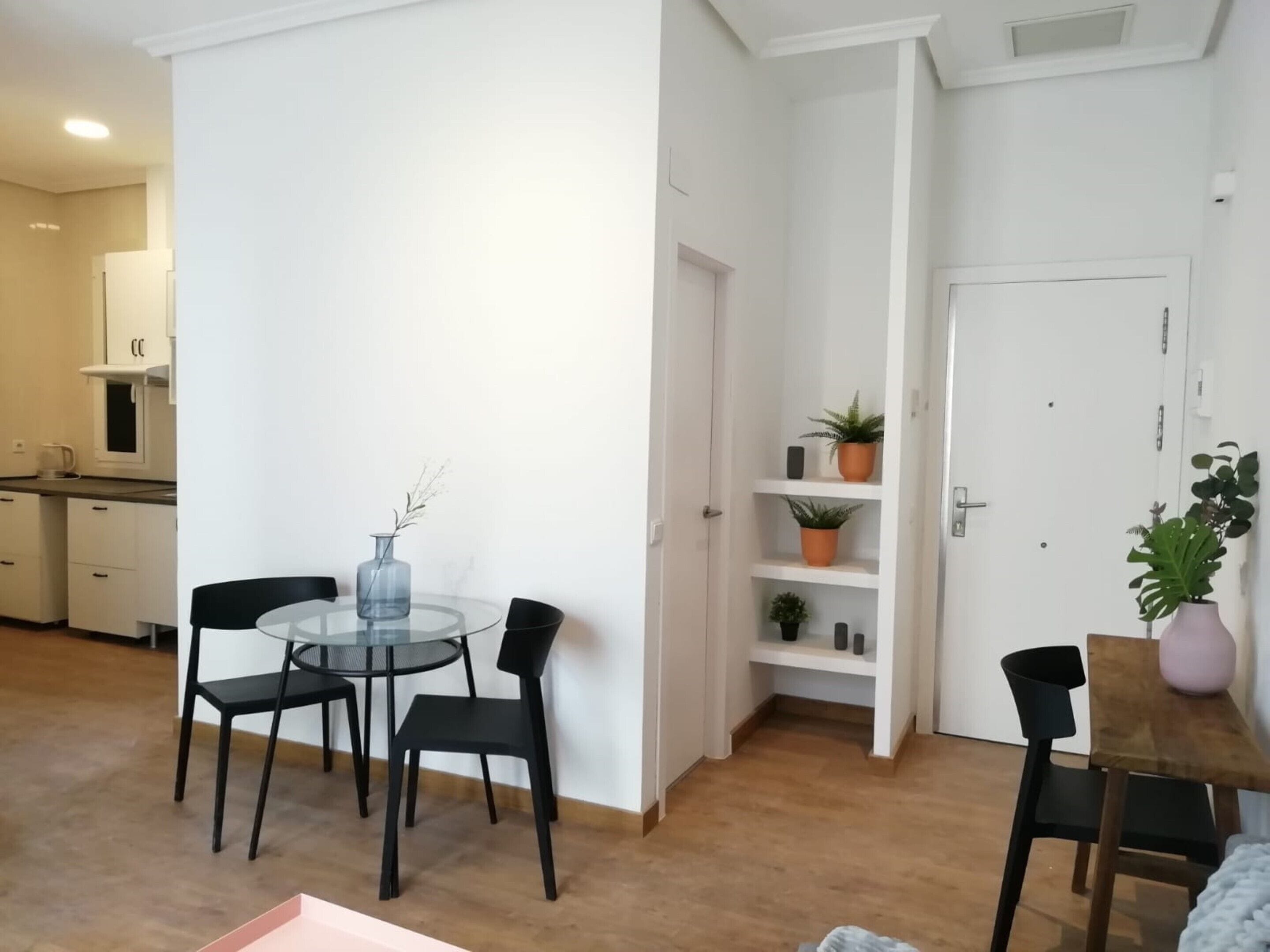 A compact living space with a round glass table and four black chairs, a wooden dining table with a single chair, a white door, and a small shelf with potted plants.
