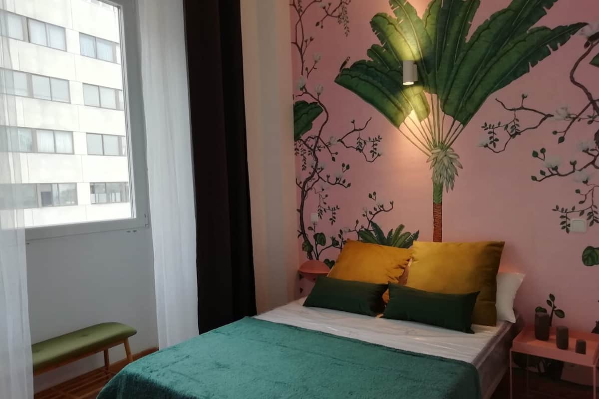 Ein Schlafzimmer mit einem Bett, grünen und gelben Kissen, einem Nachttisch, einer Bank und einem Fenster mit Vorhängen.