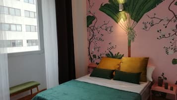 Un dormitorio con cama, almohadas verdes y amarillas, una mesita de noche, un banco y una ventana con cortinas.