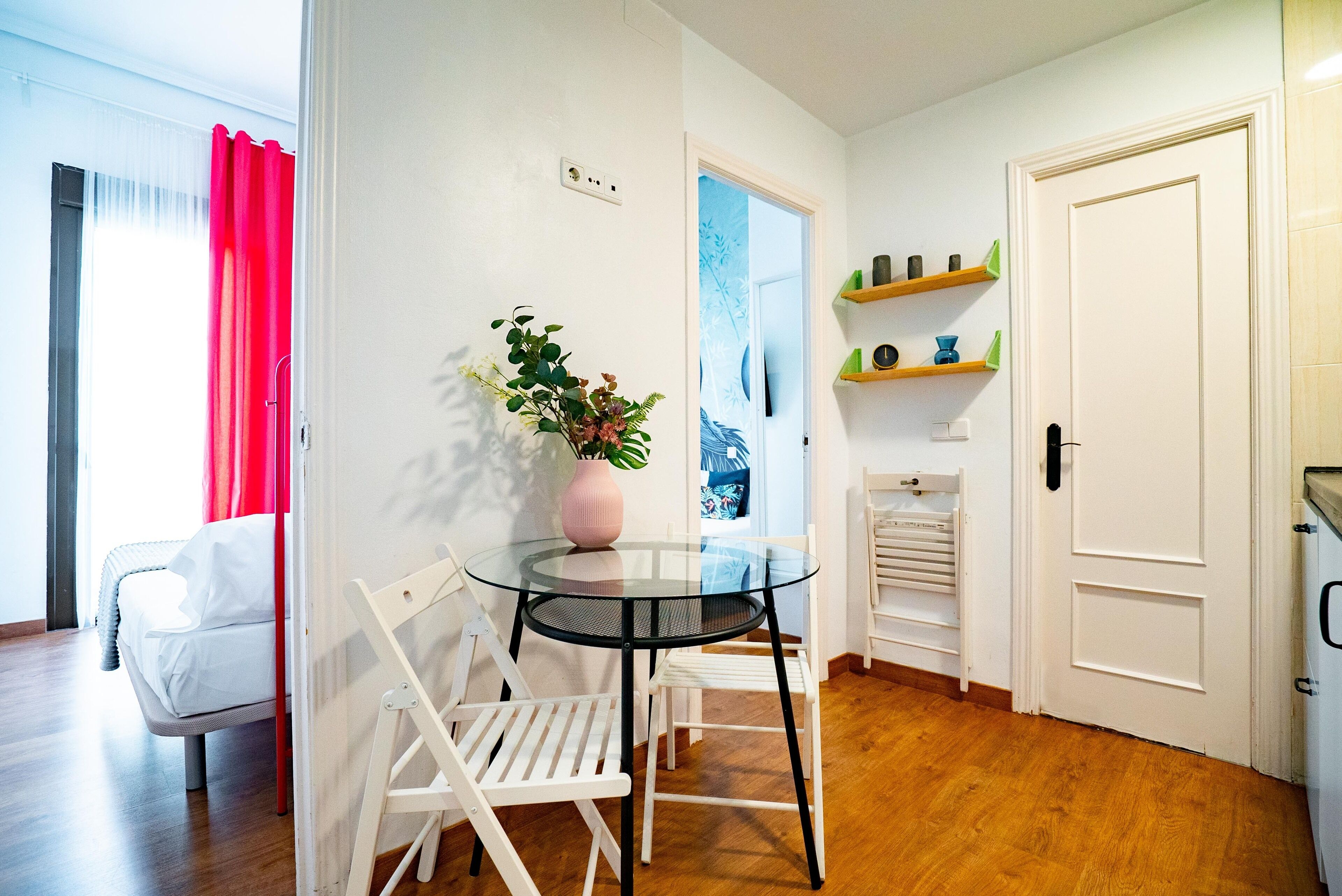 Una habitación con una mesa de vidrio, sillas blancas, un jarrón rosa con plantas, una cama con ropa de cama blanca y una puerta con estantes encima.