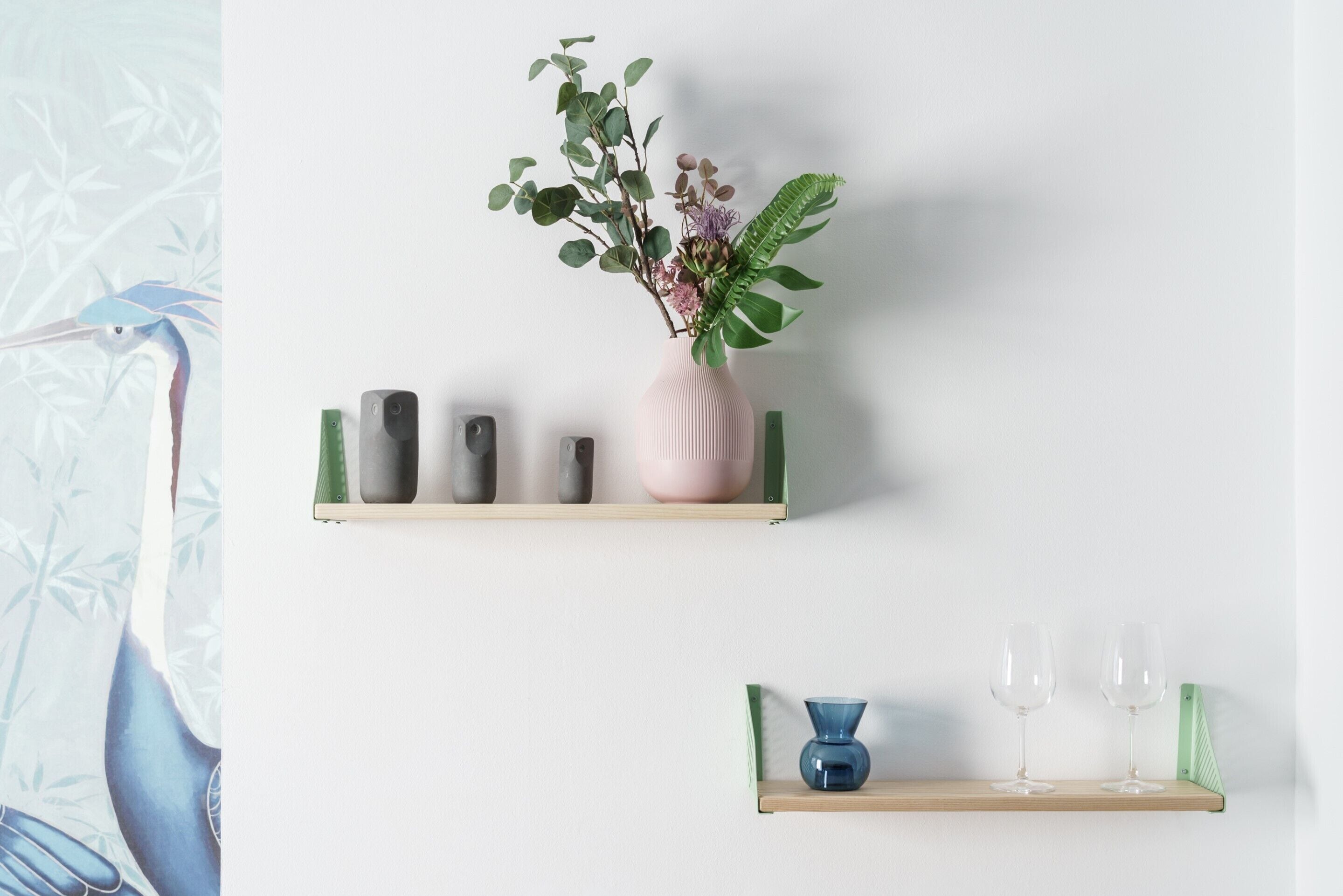 Un mur avec deux étagères ; l’étagère supérieure contient un vase avec des fleurs et des objets décoratifs, et l’étagère inférieure abrite un vase bleu et deux verres à vin.