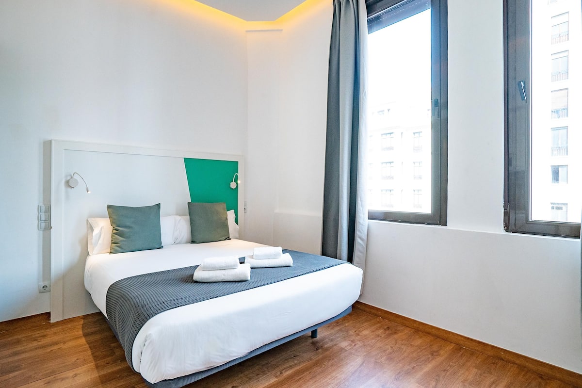 Ein ordentlich bezogenes Bett mit weißen Leinen und blauem Akzent, eine Wand in Grün und ein Fenster mit Vorhängen.