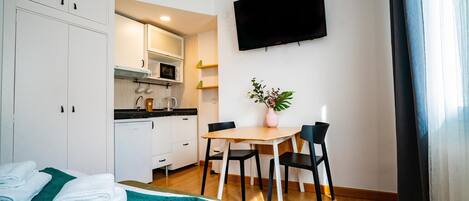 A compact hotel room with a bed, a small kitchenette, a dining table with chairs, and a TV mounted on the wall.