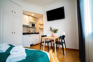 Una camera d'albergo compatta con un letto, un angolo cottura, un tavolo con sedie e un televisore fissato alla parete.