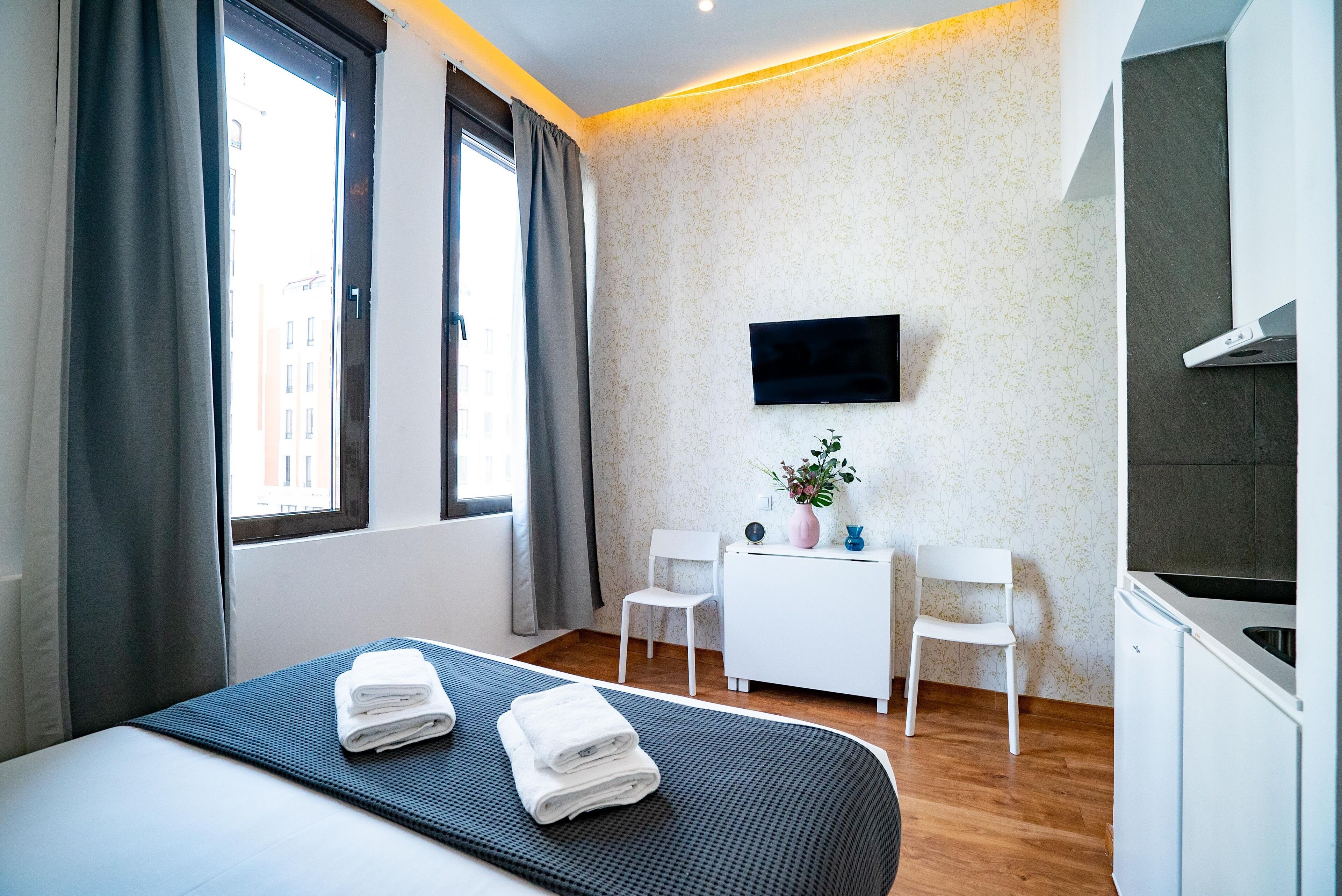 A modern hotel room with a bed, a desk, two chairs, a TV, and a kitchenette.
