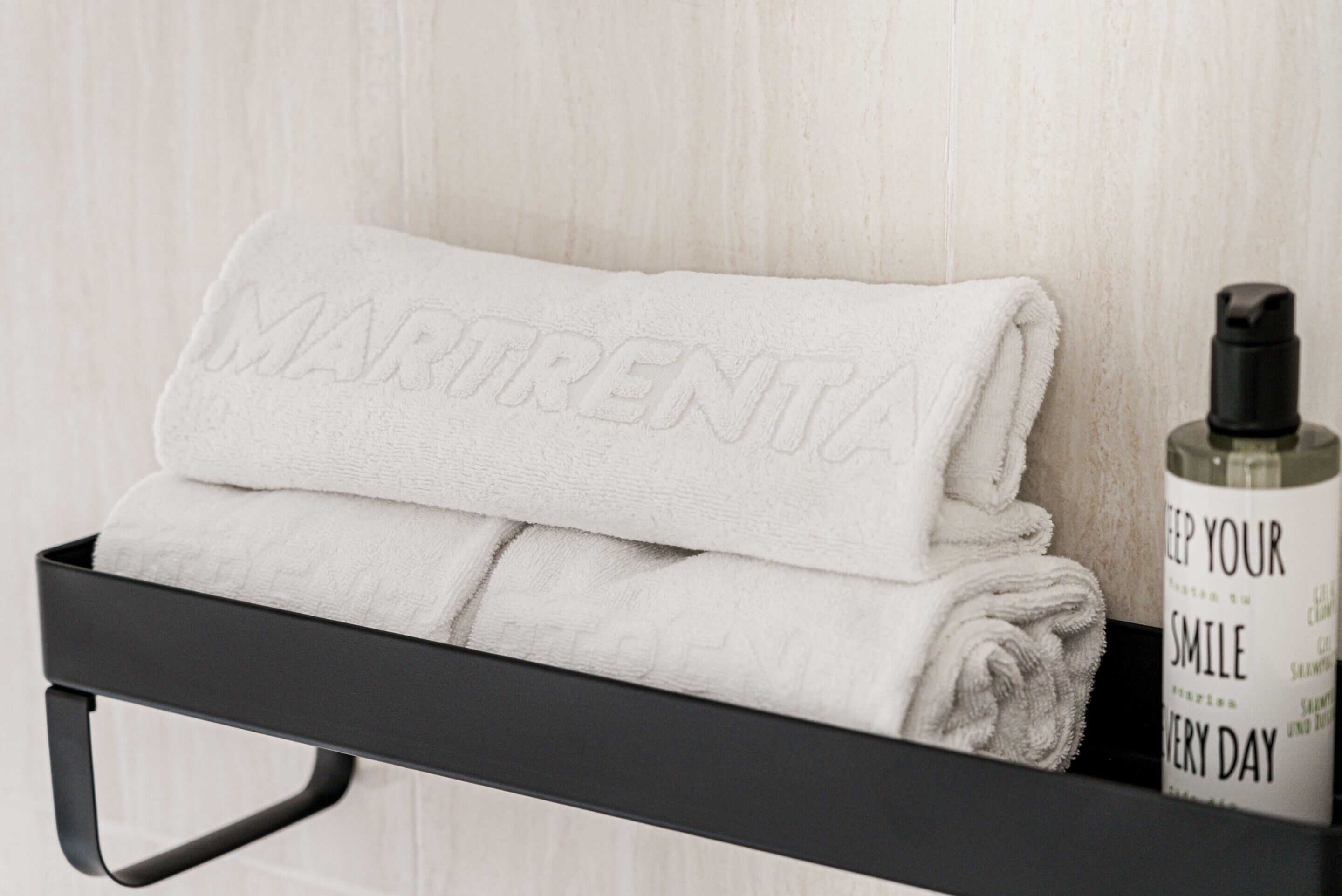 A neatly folded white towel with "MARITRENT" embossed on it, placed on a black shelf next to a bottle of shampoo.