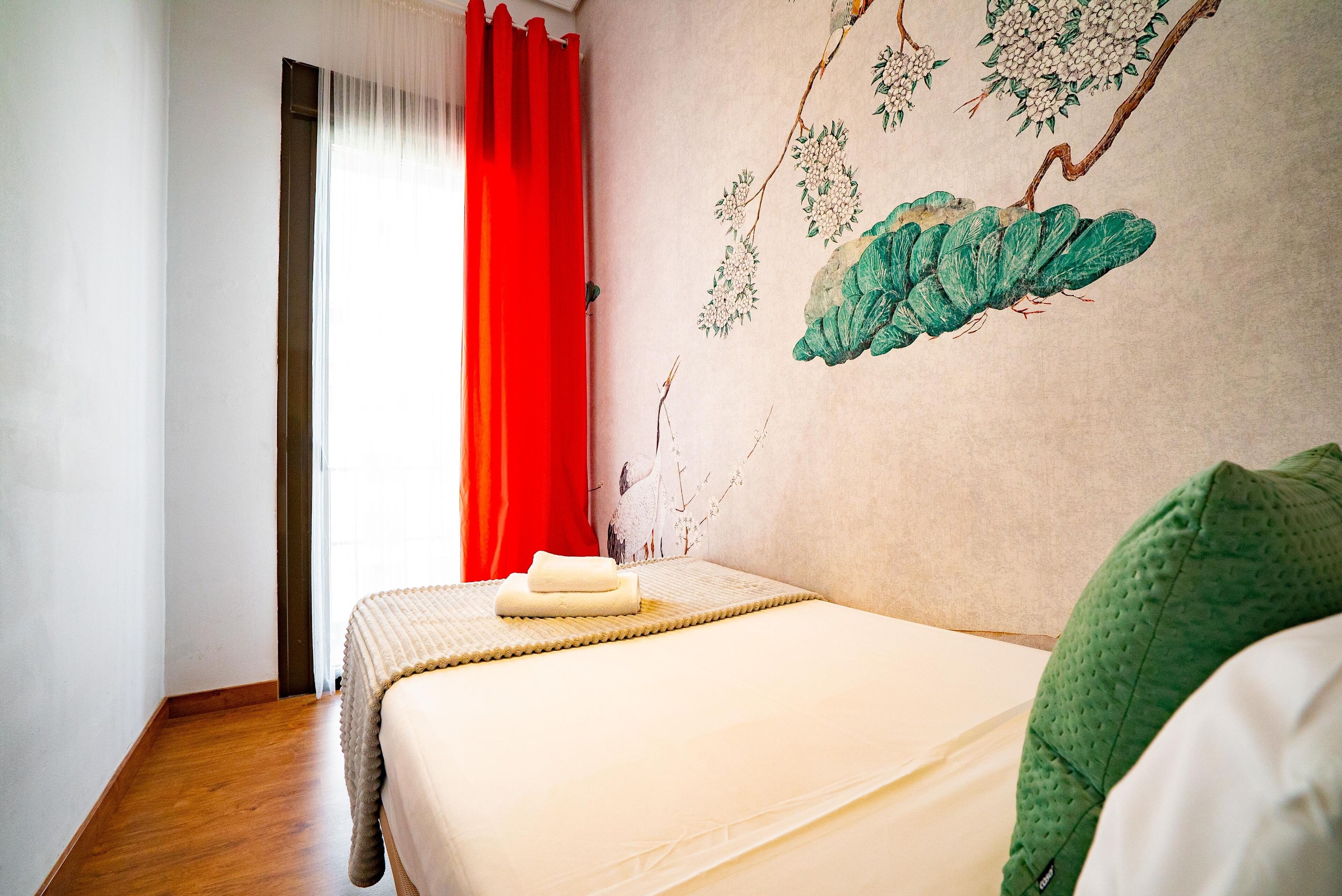 Une chambre à coucher avec un lit, un rideau rouge et un mur orné d’une fresque florale.