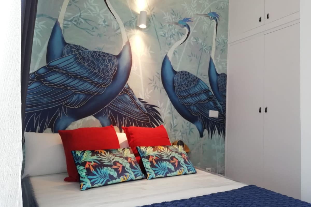 Ein Schlafzimmer mit Wandgemälde von zwei blauen Vögeln, einem Bett mit roten und farbenfrohen Kissen und einer blauen Decke.