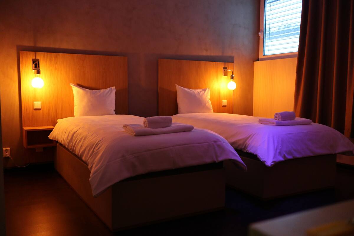 Quarto duplo com duas camas, cada uma com mesa de cabeceira, luminária na parede e janela com cortinas.
