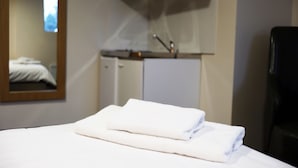 Una cama bien hecha con una toalla blanca doblada, una pequeña kitchenette con microondas y un lavabo, y un espejo que refleja parte de la habitación.