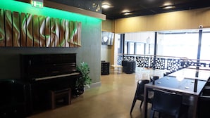 Una habitación amplia con un piano, una mesa grande y sillas. Se observa una obra de arte con iluminación verde y un televisor en la pared.