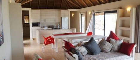 A spacious living area with a high ceiling, a sofa with red and grey cushions, a dining table, and a kitchen in the background.