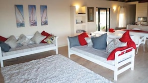 Un salon moderne avec des canapés blancs, des coussins rouges et gris, un tapis blanc et un coin repas comprenant une table et des chaises blanches.