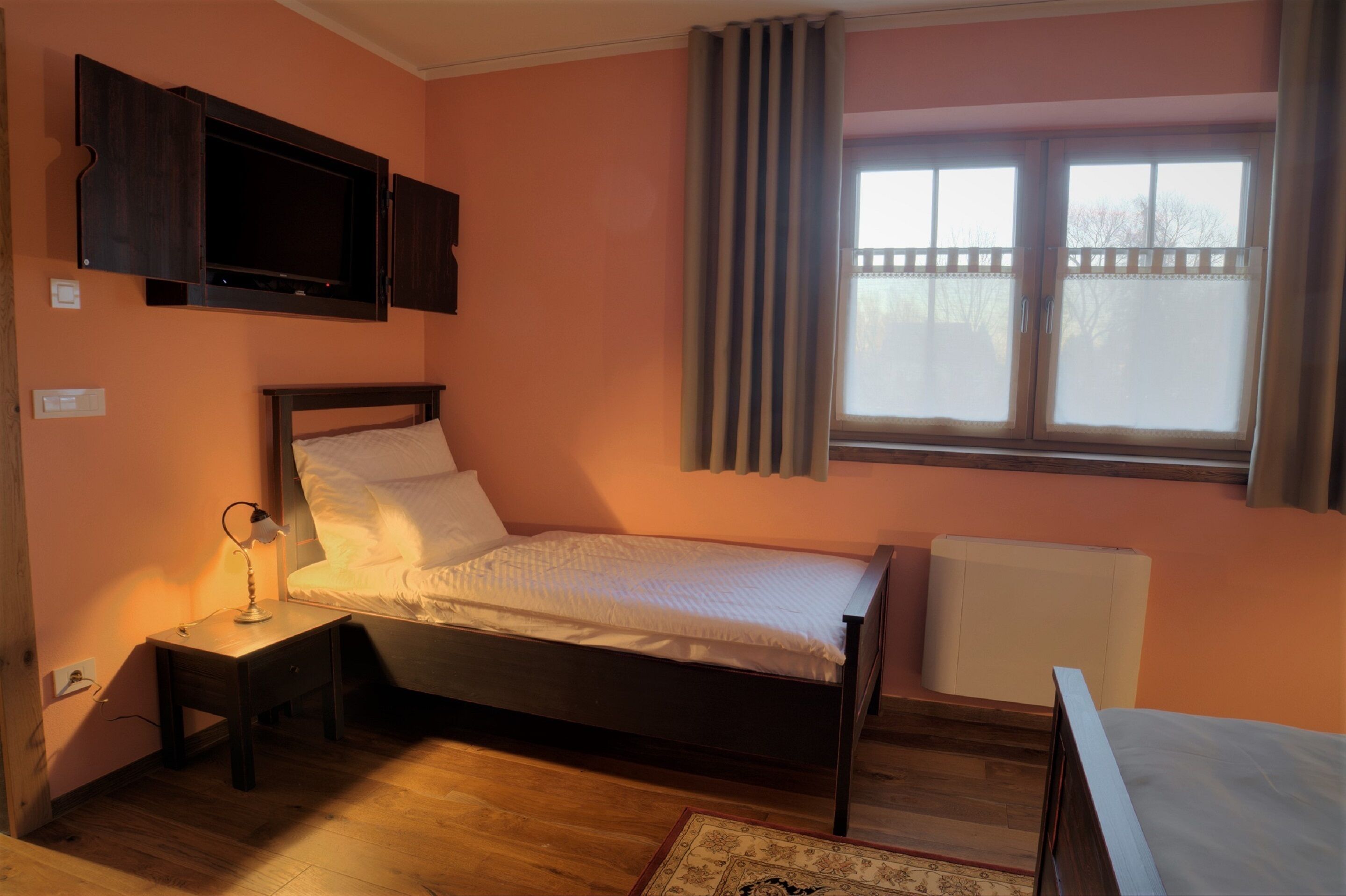 Un dormitorio con cama, mesita de noche, lámpara, televisor, ventana y radiador.