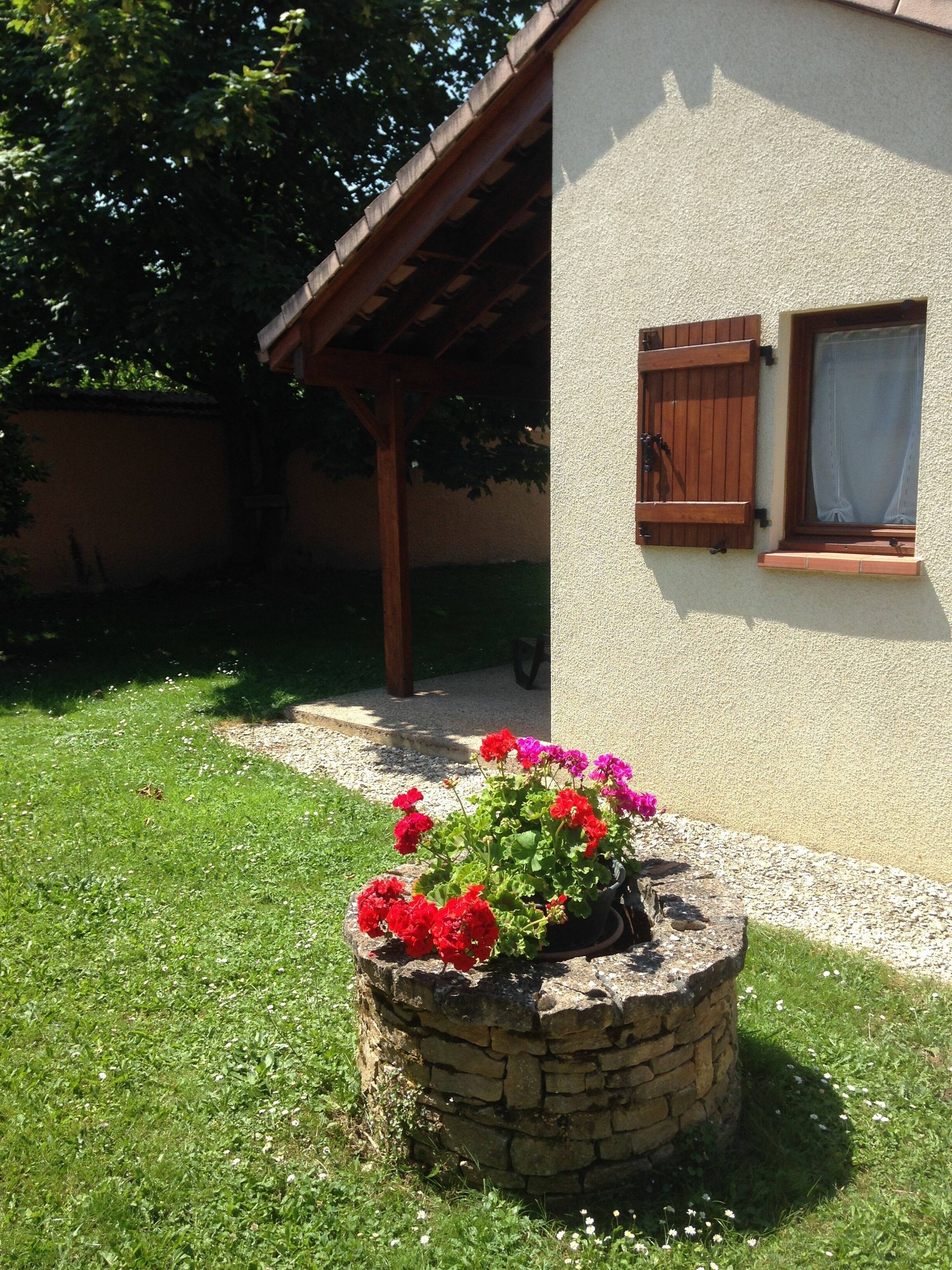 Una jardinera de piedra con flores rojas y rosas frente a una casa con contraventanas de madera y un sendero de grava.