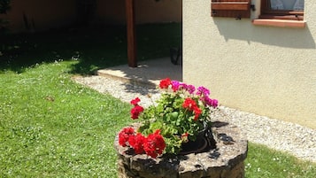 Una jardinera de piedra con flores rojas y rosas frente a una casa con contraventanas de madera y un sendero de grava.