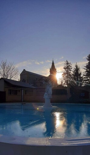 A swimming pool with a statue in the center, surrounded by buildings and trees. - Jardins du Castel Gîtes Annick et J-Paul (Saint-Georges-d'Espéranche)