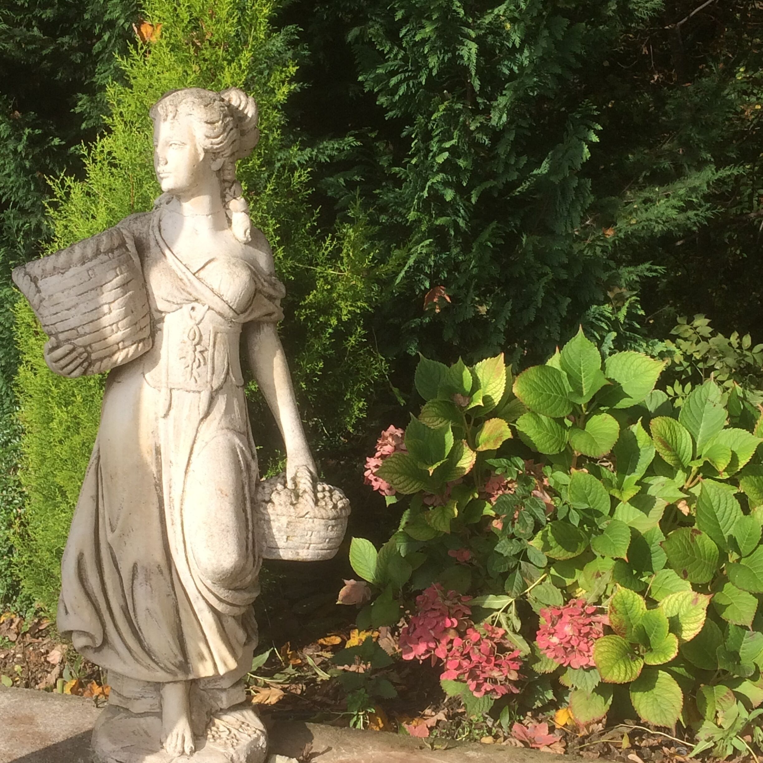 En statue af en figur der holder en kurv og en rulle, foran et baggrund af frodigt grønt og blomstrende planter.