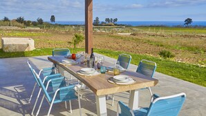 Un área de comedor al aire libre con una mesa de madera dispuesta para cuatro personas, rodeada de sillas azules, con vistas a un paisaje costero.