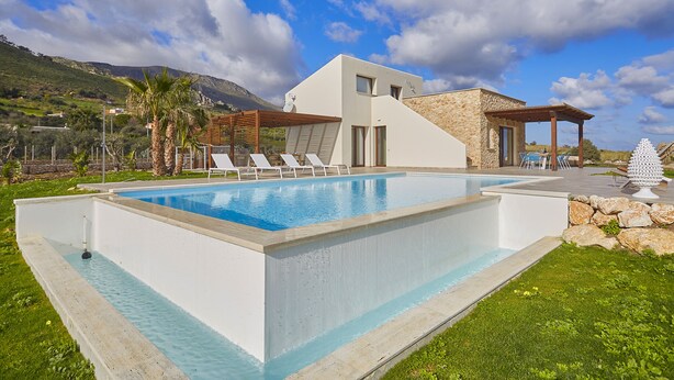 Un área de piscina moderna con una piscina rectangular, sillas de descanso y un edificio de piedra con una pérgola de madera.