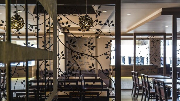 Interior de un restaurante con diseño moderno, que presenta una zona de comedor central con mesas y sillas, y detalles decorativos de metal en las paredes.