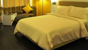 Una cama bien hecha con un edredón blanco y almohadas, un suelo de madera oscura, una mesita de noche blanca pequeña y una silla con un cojín amarillo en una habitación de hotel.