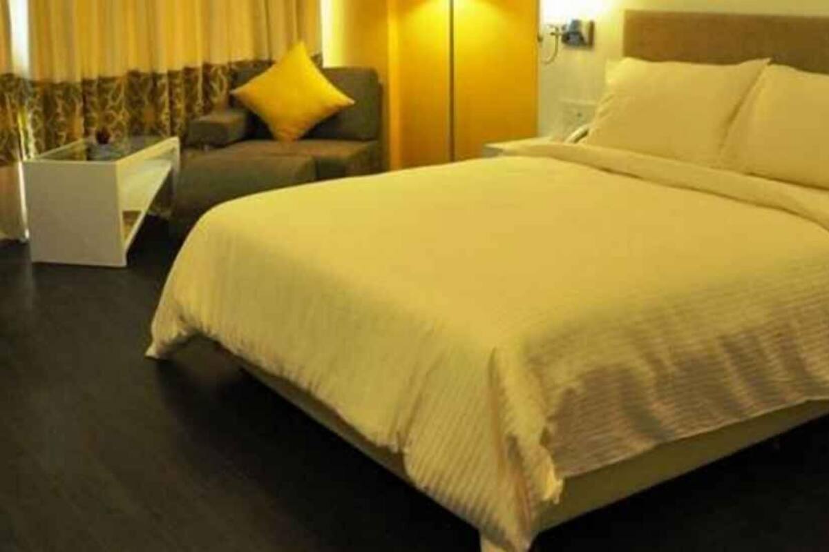 Una cama bien hecha con un edredón blanco y almohadas, un suelo de madera oscura, una mesita de noche blanca pequeña y una silla con un cojín amarillo en una habitación de hotel.