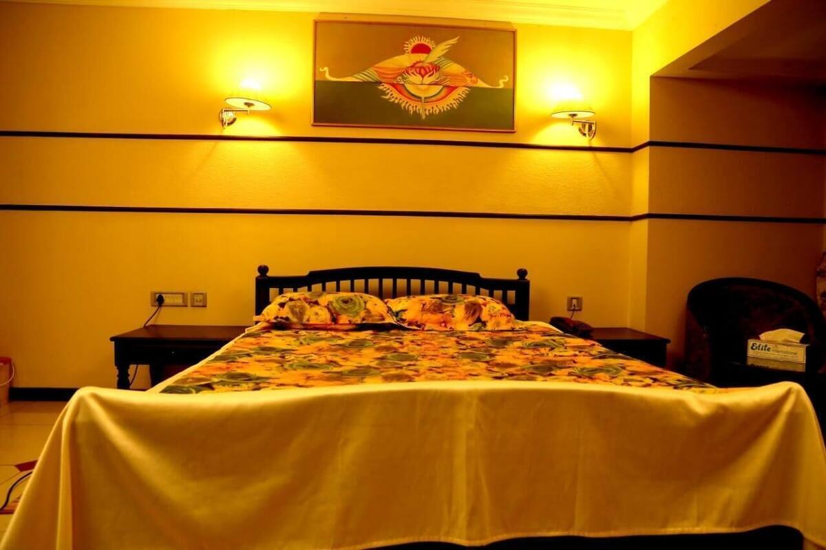 Habitación de hotel con una cama, dos lámparas fijadas a la pared, un cuadro sobre la cama y una mesita con una caja etiquetada “Elite”.