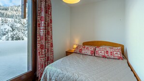 Un dormitorio con cama, cabecera de madera, almohadas estampadas, lámpara de noche y vista a un paisaje nevado a través de una ventana.