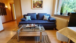 A modern living room with a dark blue sofa, a glass coffee table, and a view of greenery through a window. - FeWo Düneneck 14- OG (3) (St. Peter-Ording)