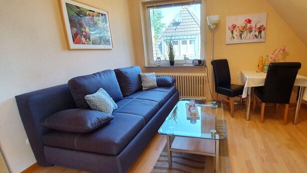 A cozy living room with a dark blue sofa, a glass coffee table, and a small dining area with a white table and black chairs. - FeWo Düneneck 14- OG (3) (St. Peter-Ording)