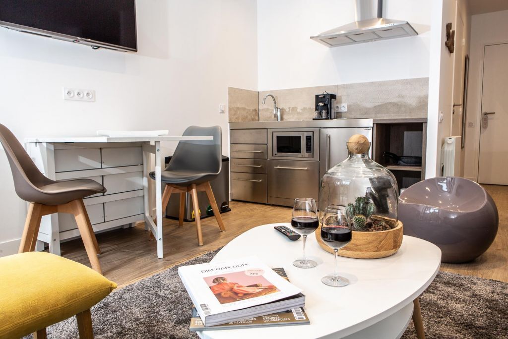 A modern living room with a white sofa, a coffee table with magazines, a TV, and a kitchen area with a stove, oven, and sink.