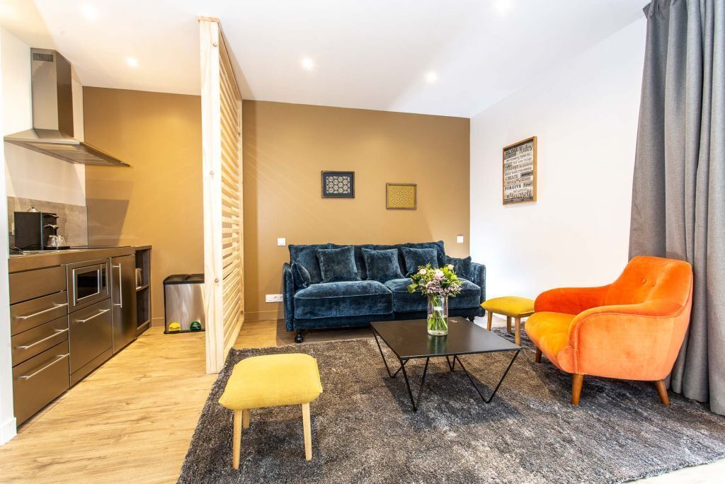 Un salon moderne avec un canapé bleu foncé, un pouf jaune et un fauteuil orange. On y trouve une table basse noire sur laquelle est posé un vase de fleurs. La pièce comprend également un coin cuisine avec des appareils en acier inoxydable et un plan de travail gris.