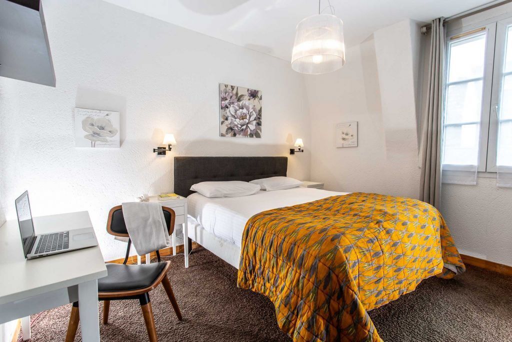 Una habitación de hotel con cama, escritorio, computadora portátil, silla y un amplio ventanal.