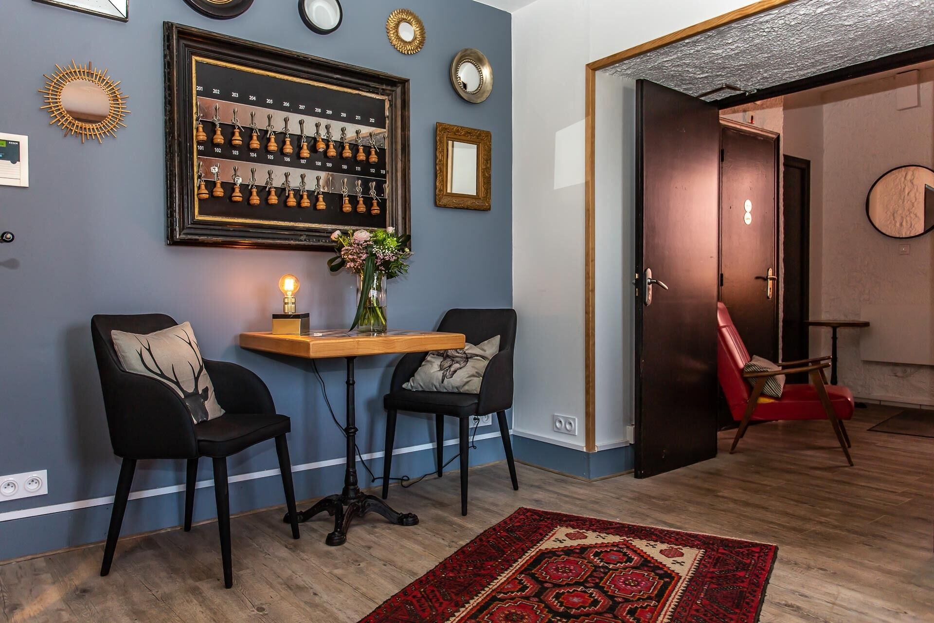 Una habitación con dos sillas negras, una pequeña mesa de madera con una lámpara, un jarrón con flores y un tablero de ajedrez en la pared.