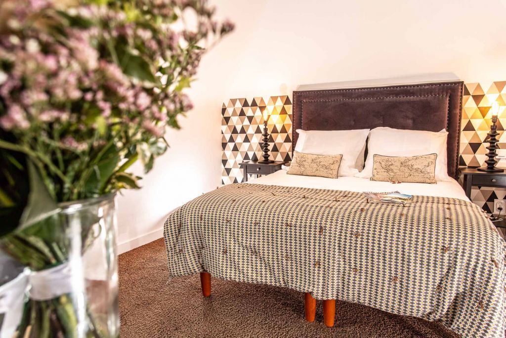 A neatly made bed with a patterned bedspread and pillows, a bedside table with a lamp, and a decorative wall with geometric patterns.