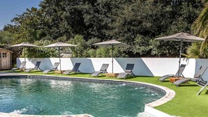 A swimming pool with a wooden deck, surrounded by lounge chairs and umbrellas, with a white wall and dense foliage in the background. - Hôtel Ty Mad (Groix)