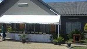 A building with a sloped roof, a covered outdoor area with a table and chairs, and a paved walkway. - Broken Hill Lodge (Kabwe)
