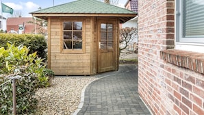 Een klein houten gebouw met een groen dak, waarschijnlijk een sauna of een klein vakantiehuisje, naast een bakstenen gebouw met een raam.
